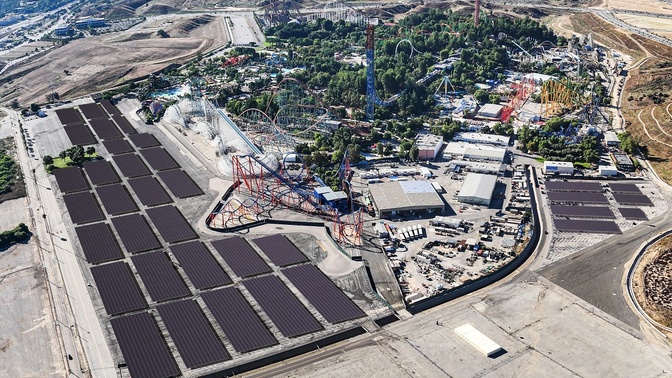 Vista aérea de um parque de diversões cercado por montanhas, com painéis solares instalados em estruturas sobre áreas de estacionamento.