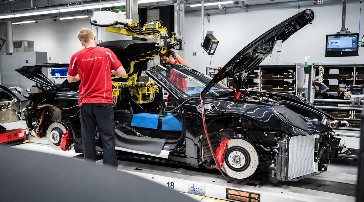 Funcionário com camiseta vermelha da Porsche trabalha ao lado de um carro esportivo preto parcialmente montado em uma linha de fábrica.