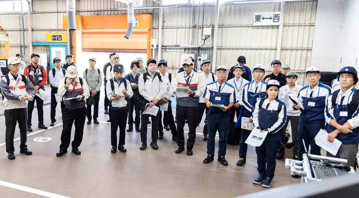 Grupo de funcionários em uniformes e bonés reunido no chão de uma fábrica; alguns seguram papéis e pranchetas.