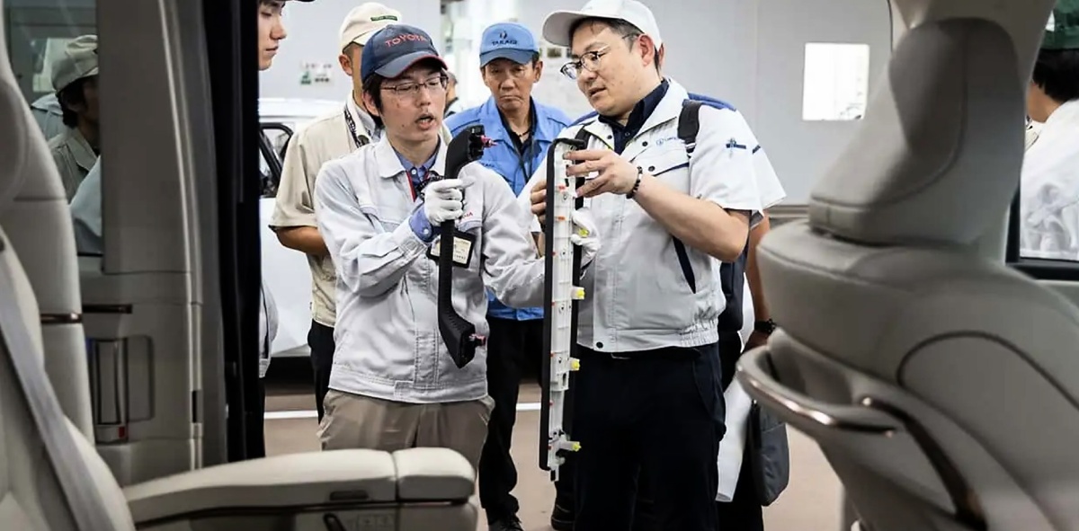 Funcionários em uniformes e bonés conversam enquanto seguram e observam uma peça automotiva comprida dentro de um veículo.