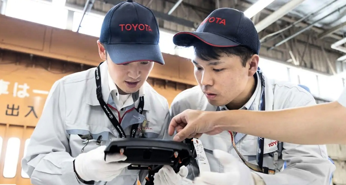 Dois funcionários com bonés da Toyota e luvas observam uma peça preta, enquanto uma mão aponta para ela.