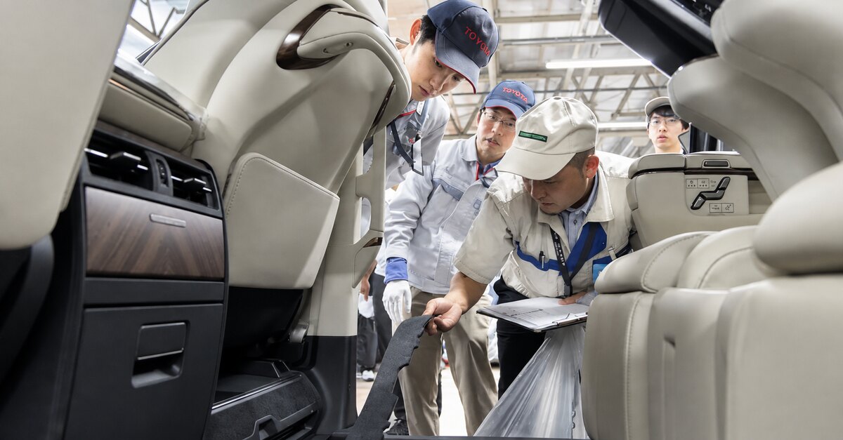 Interior bege de um veículo, com painel e bancos; funcionários com uniformes e bonés da Toyota observam uma peça escura.