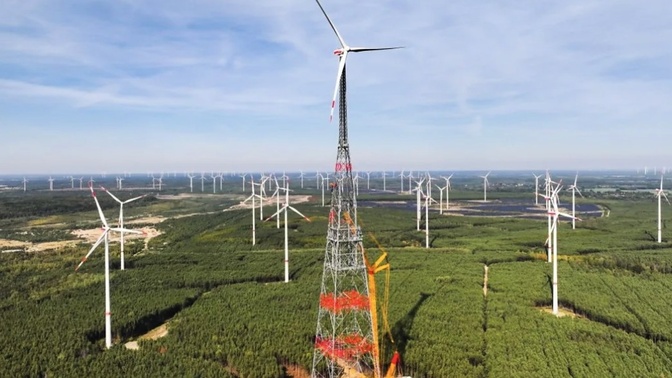 Turbina eólica de torre treliçada muito alta, com pás brancas e marcas vermelhas, entre outras turbinas sobre área florestal.
