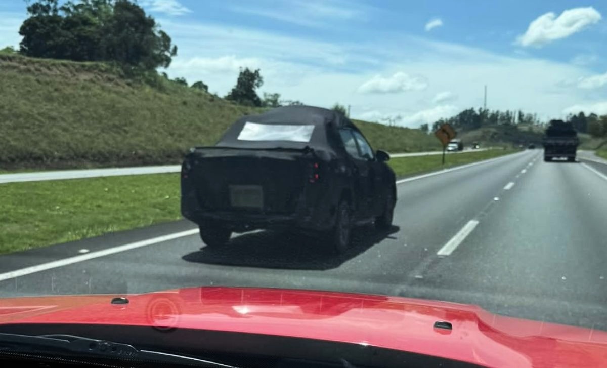 Picape Volkswagen Tukan preta e camuflada vista à distância em uma rodovia; parte do capô vermelho de outro veículo aparece em primeiro plano.