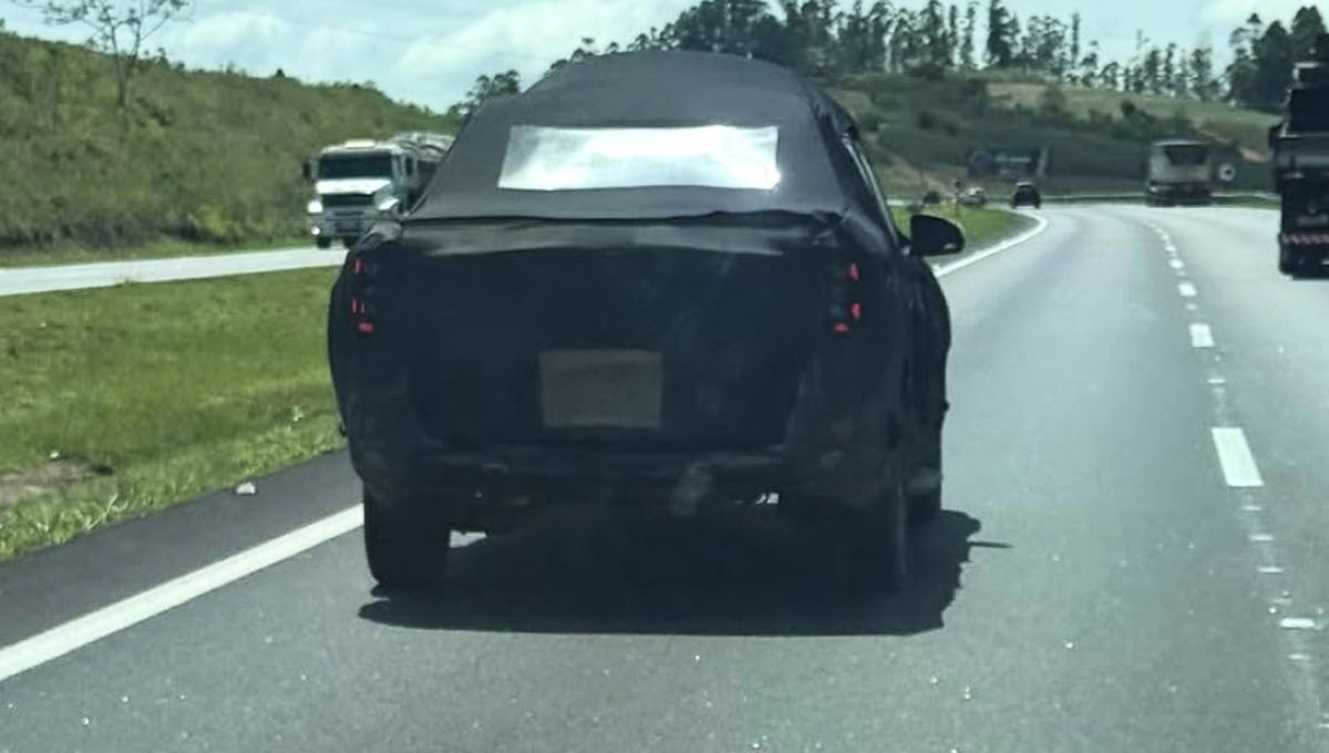 Picape Volkswagen Tukan preta e camuflada vista pela traseira, em uma rodovia com caminhões ao fundo.