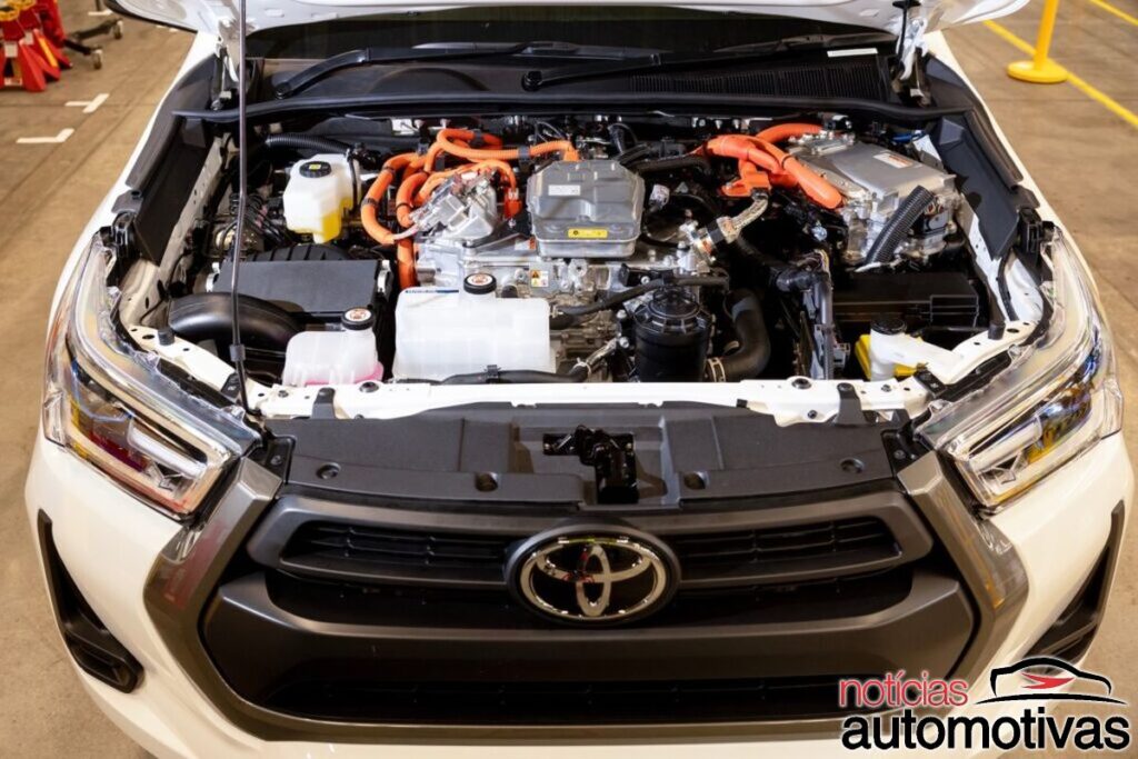 Compartimento dianteiro aberto de uma Toyota branca, com componentes mecânicos e cabos laranja visíveis.